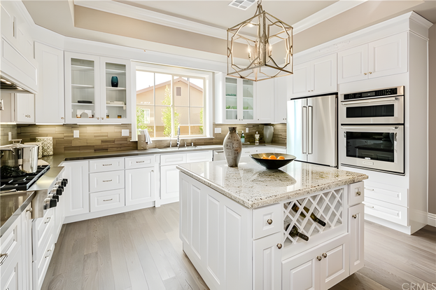 Airy kitchen layout featuring Snow Bay cabinetry, open wooden shelving, marble backsplash, and a large granite-top island.